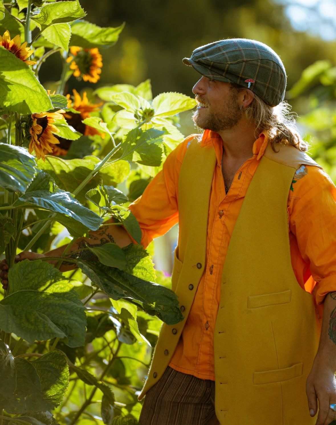 Willem Ball standing among sunflowers in an orange shirt and mustard waistcoat, looking off to one side.